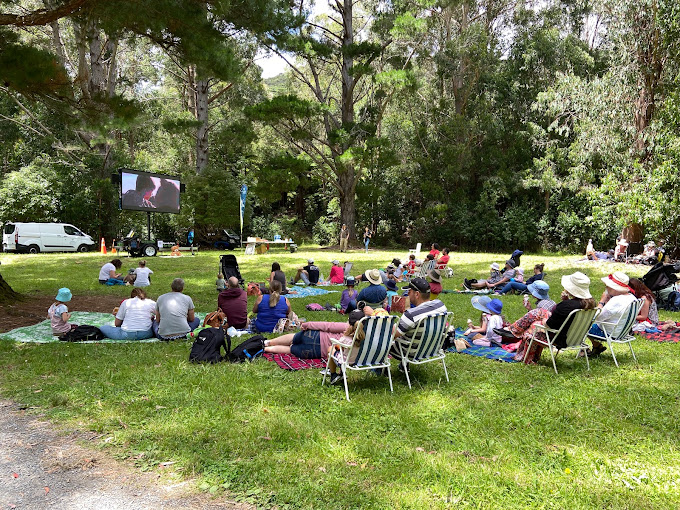A group of adults and children sit on picnic blankets watching a large screen set up in a sunny park